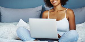 Woman of collor sitting in bed smiling with her laptop wearing blue pijama pants and a white top