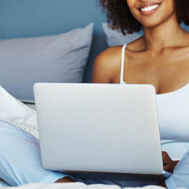 Woman of collor sitting in bed smiling with her laptop wearing blue pijama pants and a white top