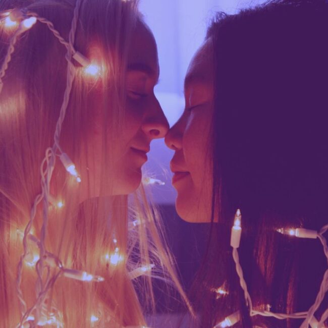 Two women about to kiss around christmas light strip