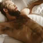 Shirtless hairy bearded man with white underwerar laying in bed surrounded by magazines