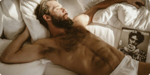 Shirtless hairy bearded man with white underwerar laying in bed surrounded by magazines