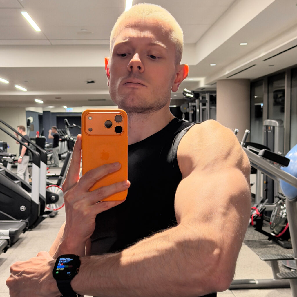 white blonde man selfie with an Iphone 17 pro max at the gym, wearing a black tank top and watch, flexing his arm