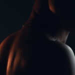 Close-up of a shirtless man’s shoulder, neck, and partially shadowed face under dramatic low lighting, creating a dark and intense mood.