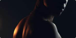 Close-up of a shirtless man’s shoulder, neck, and partially shadowed face under dramatic low lighting, creating a dark and intense mood.
