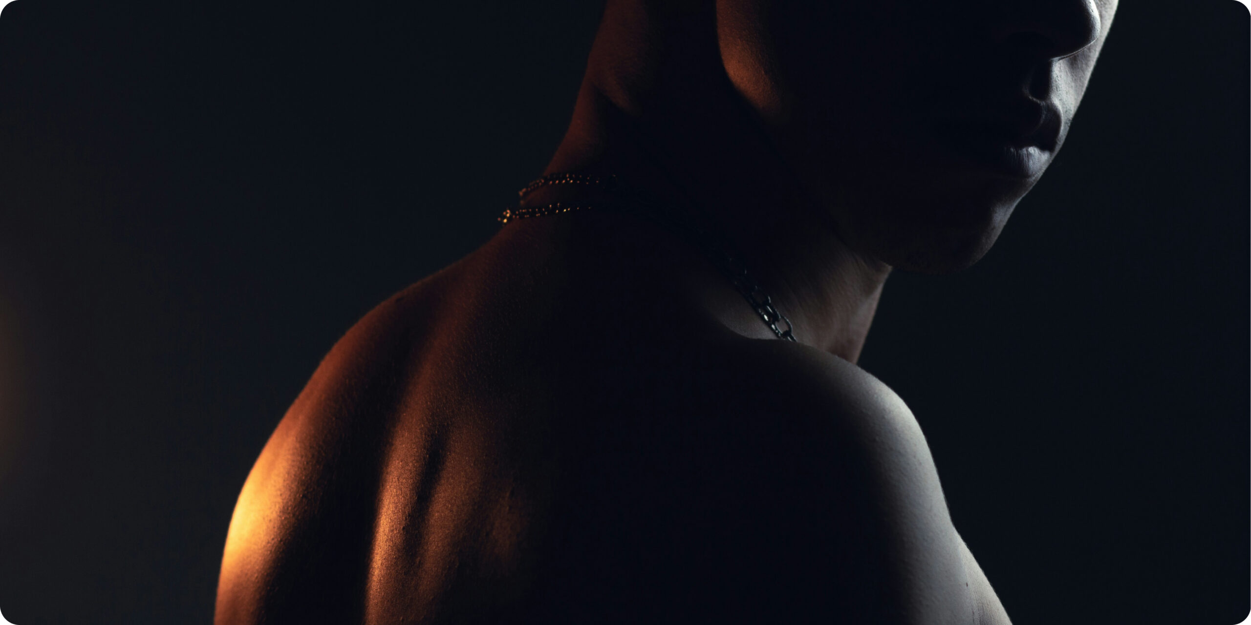 Close-up of a shirtless man’s shoulder, neck, and partially shadowed face under dramatic low lighting, creating a dark and intense mood.
