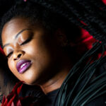 A Black woman reclining on red fabric with her eyes closed, wearing black lace clothing and holding a black-and-red feather fan in a dramatic, sensual portrait.