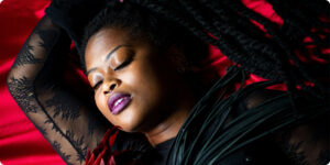 A Black woman reclining on red fabric with her eyes closed, wearing black lace clothing and holding a black-and-red feather fan in a dramatic, sensual portrait.