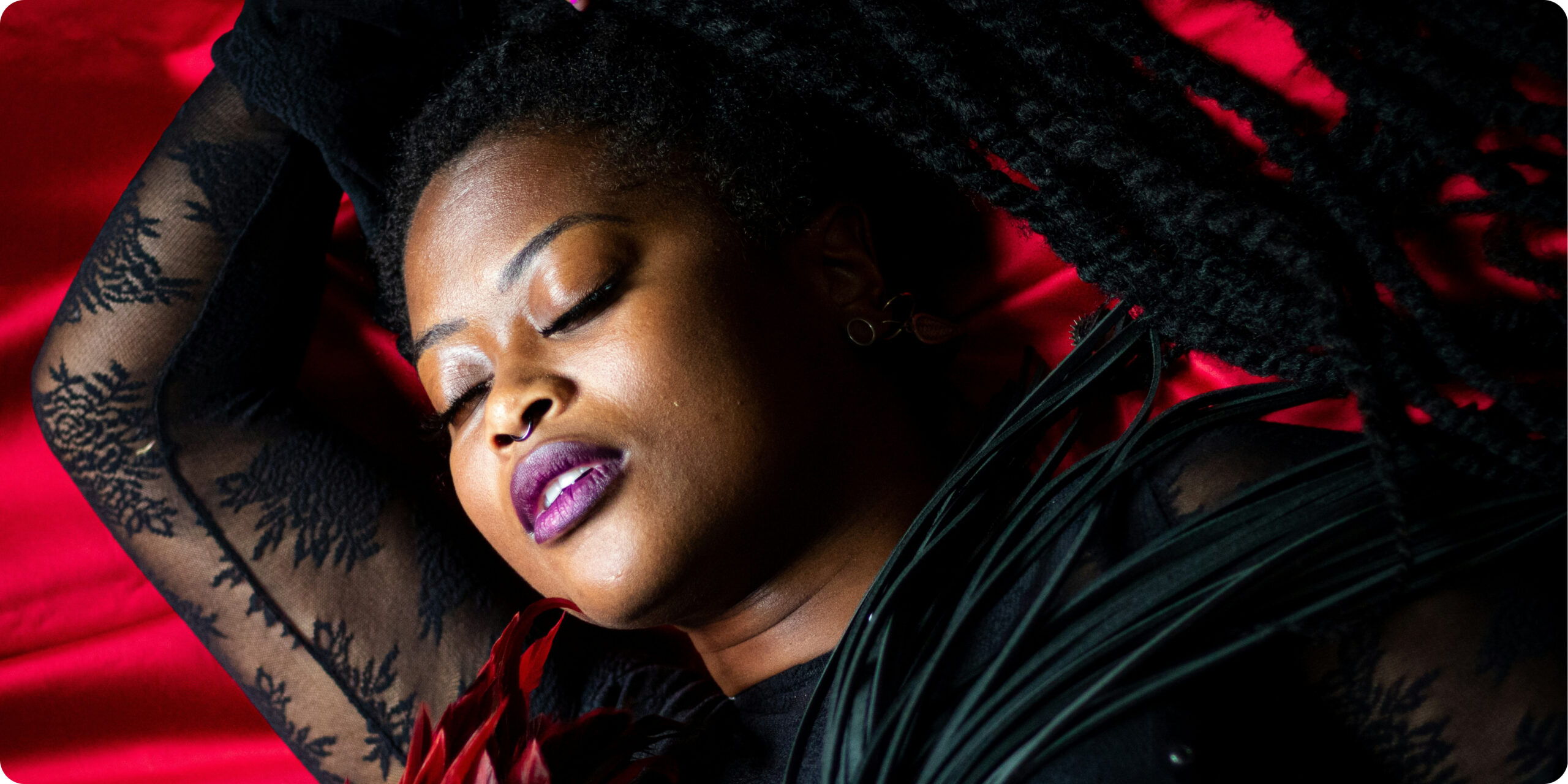 A Black woman reclining on red fabric with her eyes closed, wearing black lace clothing and holding a black-and-red feather fan in a dramatic, sensual portrait.