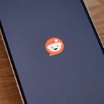 Smartphone displaying the Reddit app logo — the Reddit alien mascot (Snoo) in orange — against a dark screen, placed on a wooden surface.