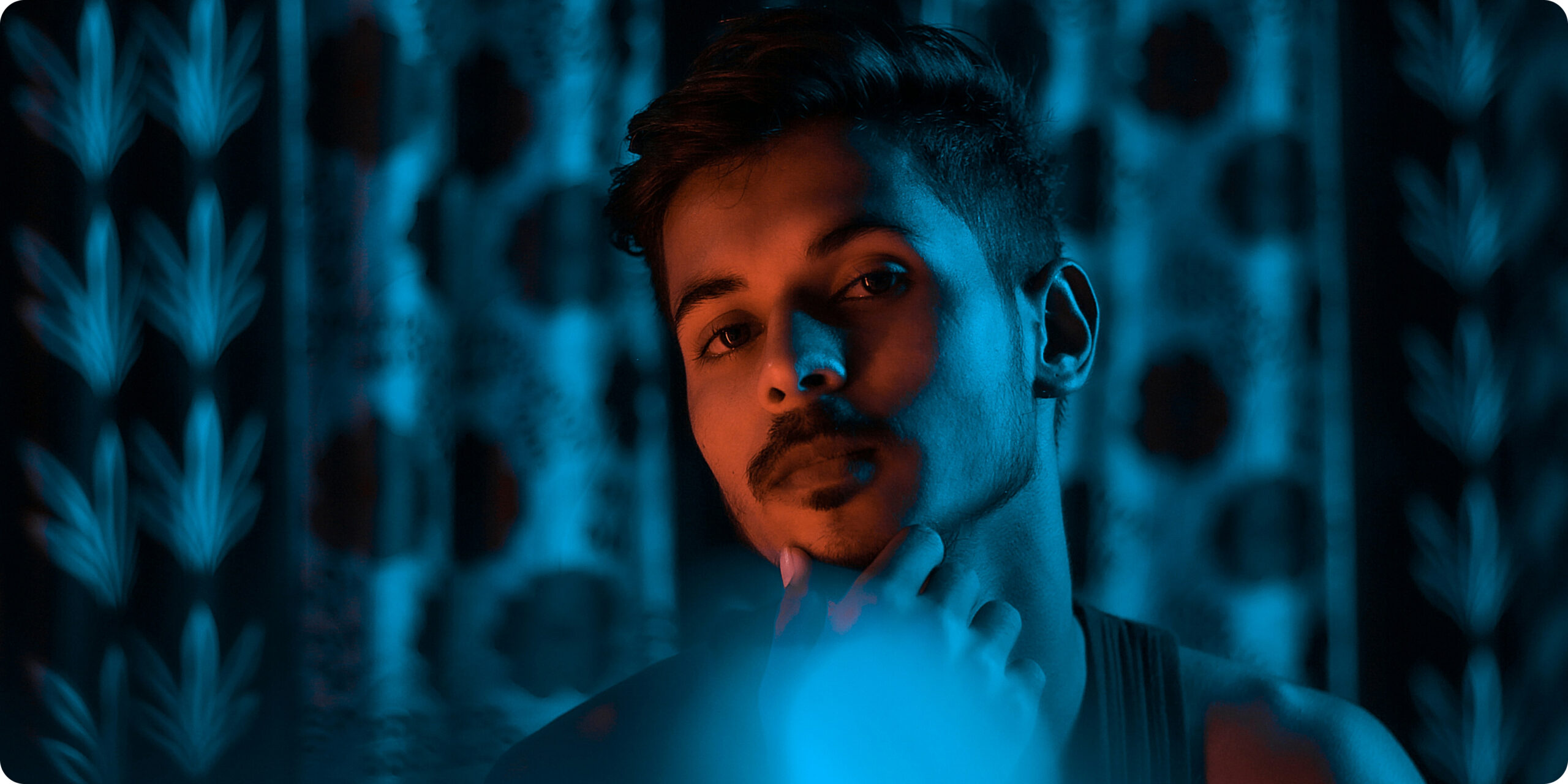 Thoughtful young man with a mustache looking into a mirror under blue lighting, symbolizing independent content creation.