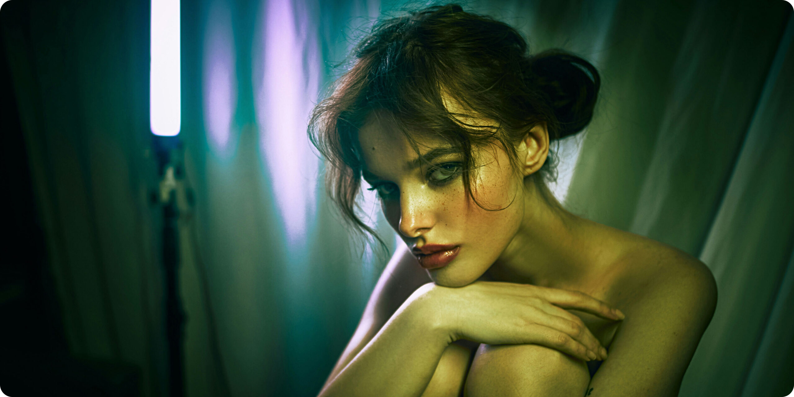A woman with dark hair and makeup sits in a dimly lit room, illuminated by green and purple lighting, looking toward the camera with her arms resting on her knees.