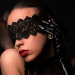A woman with dark slicked-back hair wears a black lace blindfold and a shiny black satin glove, posed against a black background with red lips slightly parted.