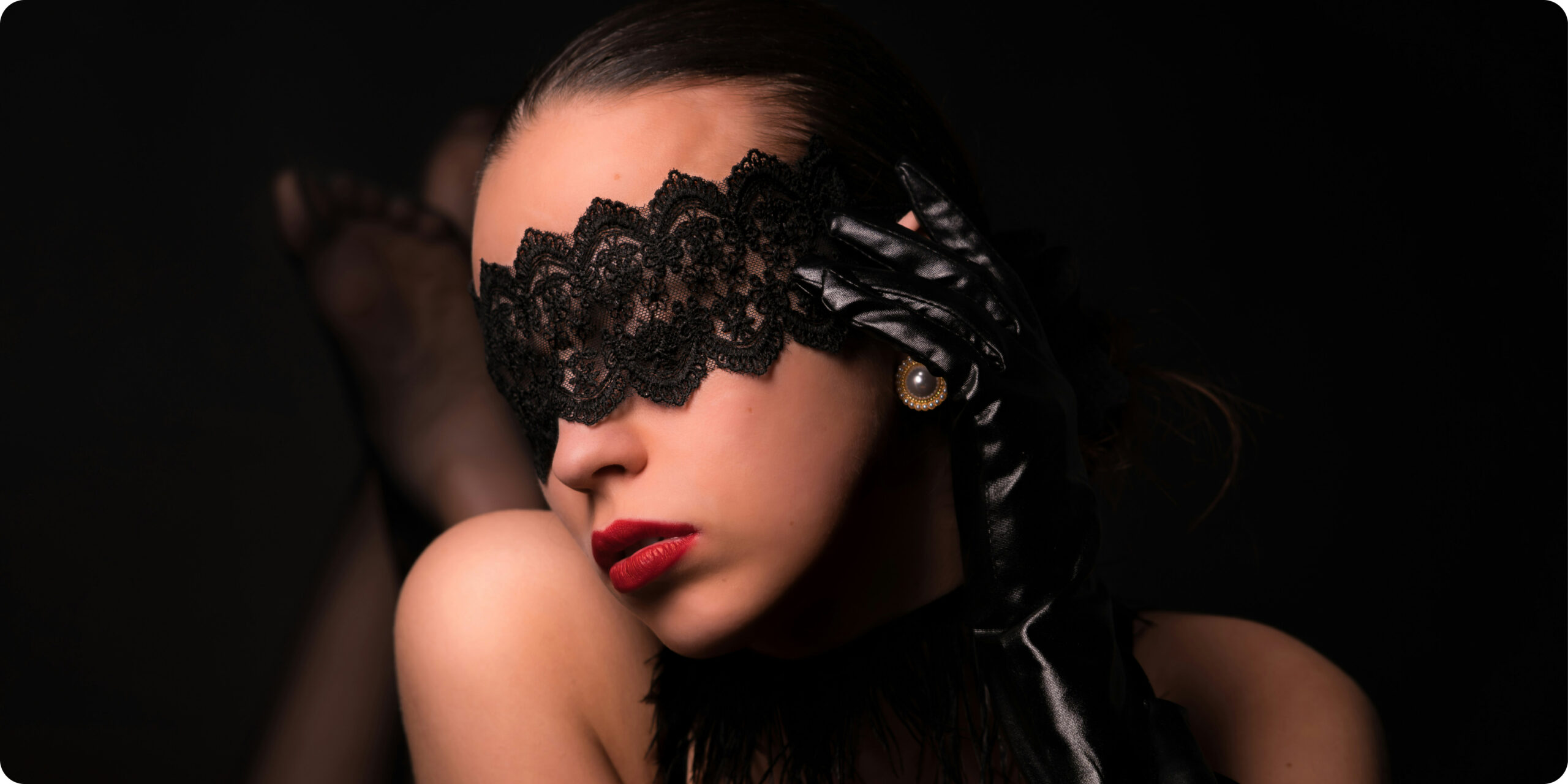A woman with dark slicked-back hair wears a black lace blindfold and a shiny black satin glove, posed against a black background with red lips slightly parted.
