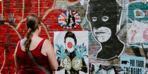 A woman with a ponytail, in a red tank top and gray crossbody bag, viewed from behind as she looks at a brick wall covered in red graffiti and street-art posters.