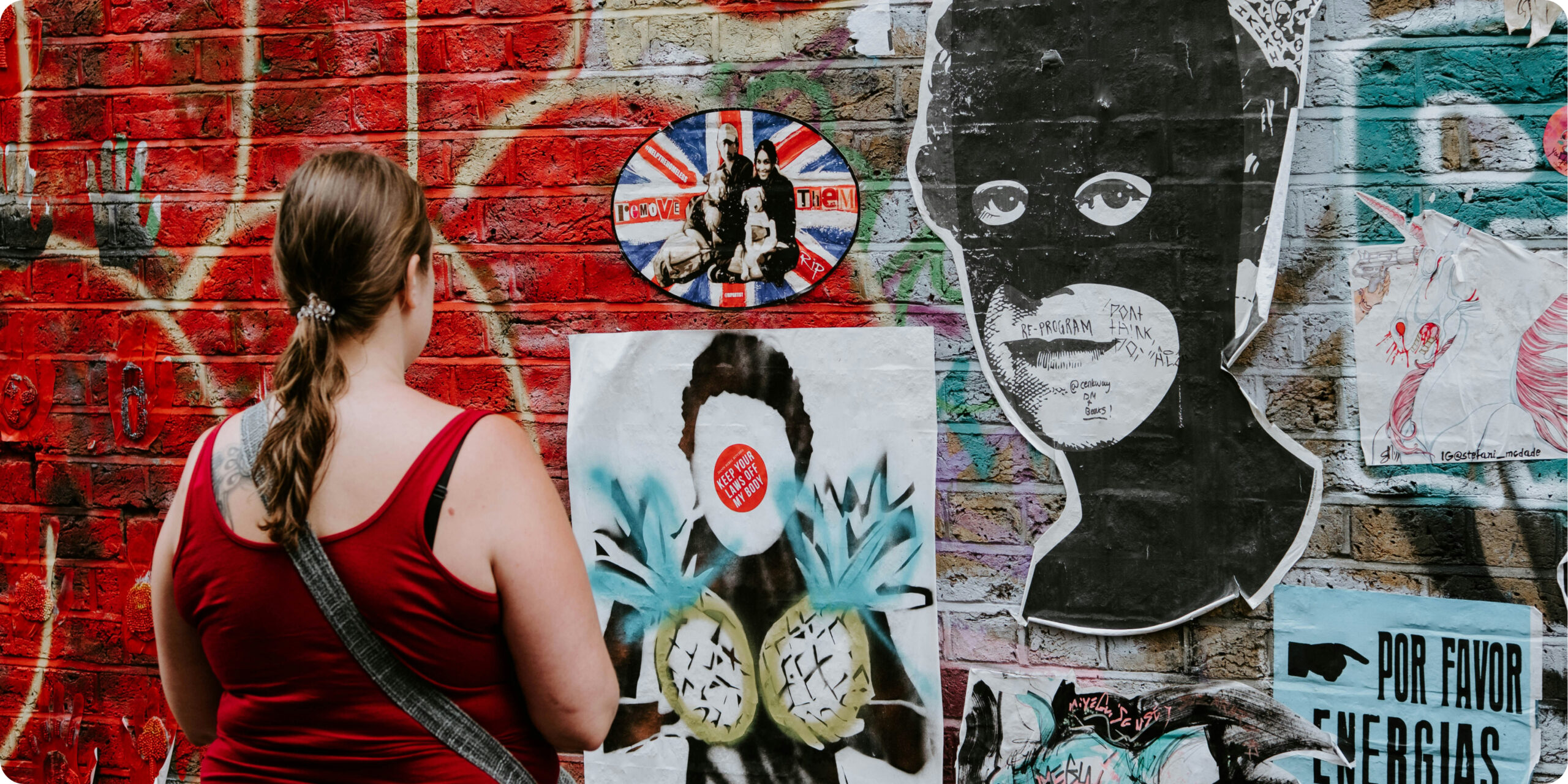 A woman with a ponytail, in a red tank top and gray crossbody bag, viewed from behind as she looks at a brick wall covered in red graffiti and street-art posters.