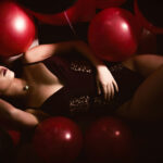 A woman in black lingerie reclines among red and black balloons in a dimly lit setting.