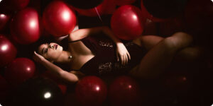 A woman in black lingerie reclines among red and black balloons in a dimly lit setting.